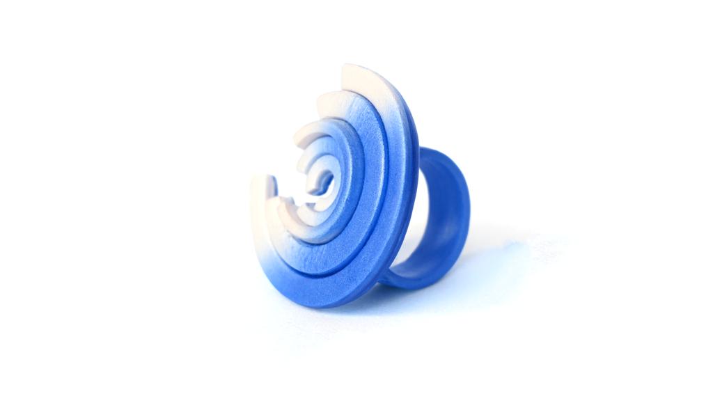 Anillo artesanal de arcilla polimérica con diseño formado por segmentos de círculos concéntricos en colores metálicos, degradados de azul a blanco.