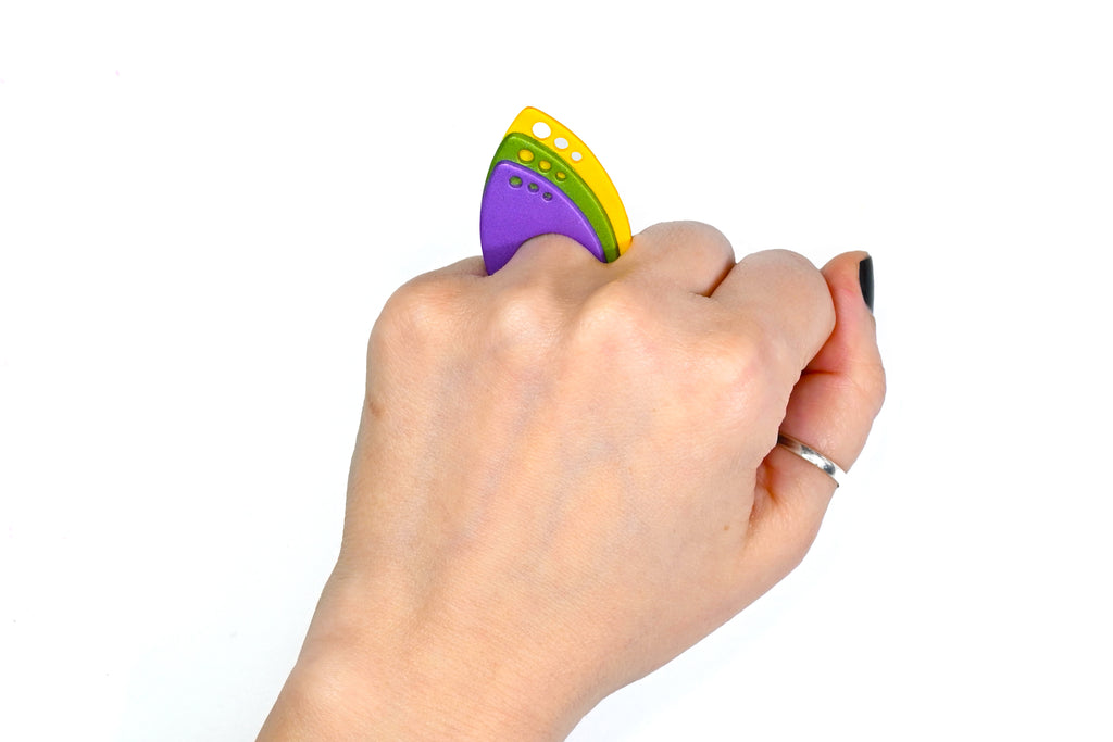 Mano luciendo el anillo artesanal de arcilla polimérica modelo Tríada, en colores púrpura, verde y amarillo.